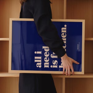 May include: A rectangular framed print with a dark blue background and white text that reads all i need is h...en. The text is arranged vertically in a bold, sans-serif font. The frame is made of light-colored wood with a smooth, natural finish.