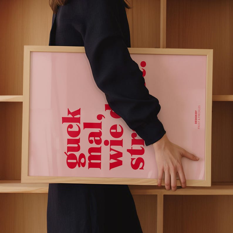 May include: A framed pink art print with red text that reads "guck mal, wie du strahlst." The print is held by a person in front of a wooden shelf. The frame is a light wood color, and the design is by Frost & Freckles.