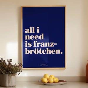 May include: A rectangular wall art print in a light wood frame featuring a deep navy blue background with the white text "all i need is franz-br&ouml;tchen." and the smaller text "DESIGN BY FROST & FRECKLES" centered at the bottom of the print.