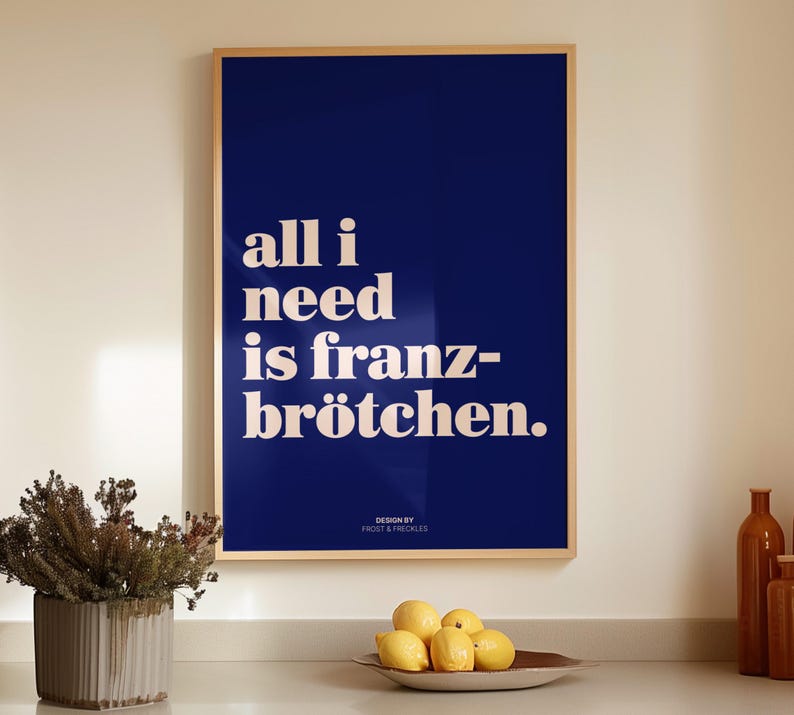 May include: A rectangular wall art print in a light wood frame featuring a deep navy blue background with the white text "all i need is franz-br&ouml;tchen." and the smaller text "DESIGN BY FROST & FRECKLES" centered at the bottom of the print.