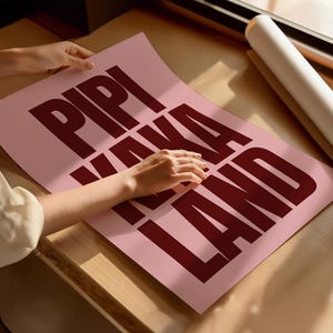 Peut inclure: Une affiche rose avec les mots "PIPI KAKA LAND" en gras, rouge fonc&eacute;. L'affiche est pos&eacute;e sur une surface en bois pr&egrave;s d'une fen&ecirc;tre. Un rouleau de papier blanc est &eacute;galement visible.