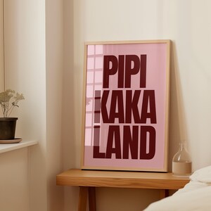 Peut inclure: Une affiche encadr&eacute;e avec les mots "PIPI KAKA LAND" en lettres majuscules rouge fonc&eacute; sur un fond rose. Le cadre est en bois clair, et l'affiche est expos&eacute;e sur une table en bois.