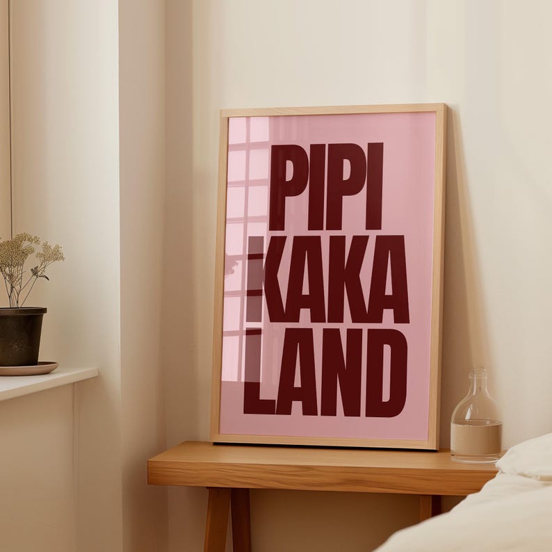 Peut inclure: Une affiche encadr&eacute;e avec les mots "PIPI KAKA LAND" en lettres majuscules rouge fonc&eacute; sur un fond rose. Le cadre est en bois clair, et l'affiche est expos&eacute;e sur une table en bois.