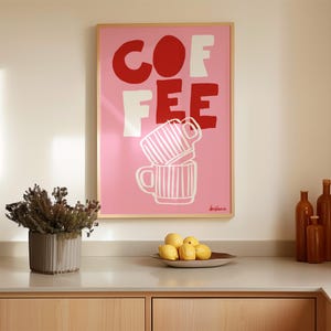 May include: A framed pink art print with the word "COFFEE" in red and white block letters. Below the text are two stacked coffee mugs with vertical stripes. The print is displayed above a countertop with lemons and decorative bottles.