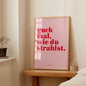 May include: A framed pink poster with the German phrase "guck mal, wie du strahlst" in red lettering. The poster is in a light wood frame and sits on a wooden table. A small glass bottle is on the table.