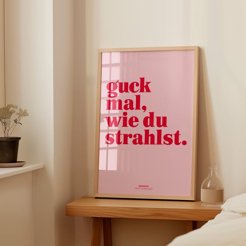 May include: A framed pink poster with the German phrase "guck mal, wie du strahlst" in red lettering. The poster is in a light wood frame and sits on a wooden table. A small glass bottle is on the table.