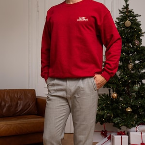 May include: A red crewneck sweatshirt featuring the text MERRY CHRISTMAS embroidered in white on the upper left chest, paired with light gray trousers. The sweatshirt has long sleeves with ribbed cuffs and a ribbed hemline, providing a classic fit.