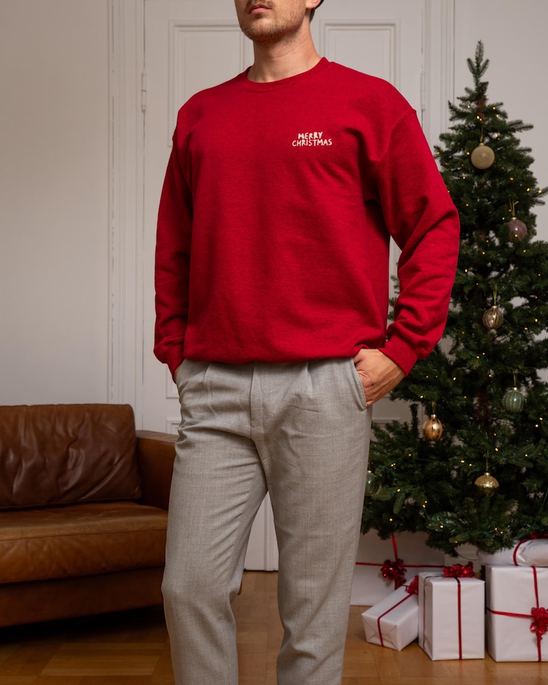 May include: A red crewneck sweatshirt featuring the text MERRY CHRISTMAS embroidered in white on the upper left chest, paired with light gray trousers. The sweatshirt has long sleeves with ribbed cuffs and a ribbed hemline, providing a classic fit.