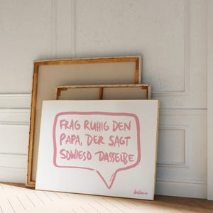 May include: A white canvas print with a pink speech bubble that says "Frag ruhig den Papa, der sagt sowieso dasselbe". The print is framed in a gold frame and is leaning against a white wall.