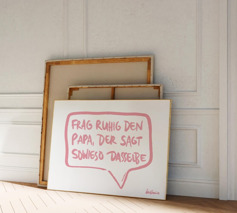 May include: A white canvas print with a pink speech bubble that says "Frag ruhig den Papa, der sagt sowieso dasselbe". The print is framed in a gold frame and is leaning against a white wall.