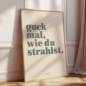 May include: A framed poster with the German phrase "guck mal, wie du strahlst" in green text on a beige background. The poster is leaning against a wall and has a wooden frame. The design is by Frost & Freckles.