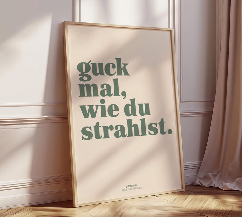 May include: A framed poster with the German phrase "guck mal, wie du strahlst" in green text on a beige background. The poster is leaning against a wall and has a wooden frame. The design is by Frost & Freckles.