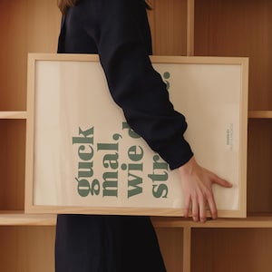 May include: A rectangular framed print with the text "guck mal, wie str..." in green, set against a cream background. The frame is made of light-colored wood. The design is by Frost & Freckles.