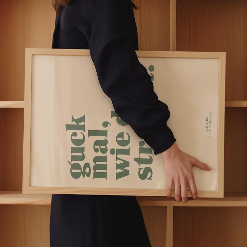 May include: A rectangular framed print with the text "guck mal, wie str..." in green, set against a cream background. The frame is made of light-colored wood. The design is by Frost & Freckles.