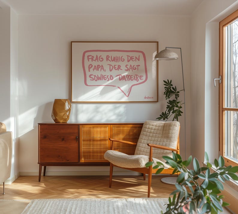 May include: A framed print with a pink speech bubble that says "Frag ruhig den Papa, der sagt sowieso dasselbe". The print is hanging on a white wall in a living room with a wooden cabinet, a chair, and a plant.