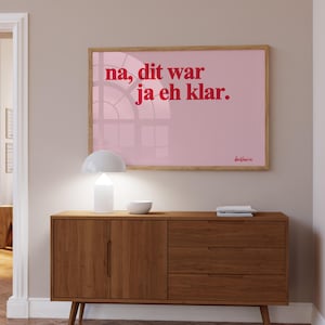 Könnte beinhalten: Gerahmter rosa Druck mit rotem Text "na, dit war ja eh klar." Über einem Holzschrank mit weißer Lampe und Schale. Der Schrank hat zwei Türen und drei Schubladen. Neutrale Inneneinrichtung.