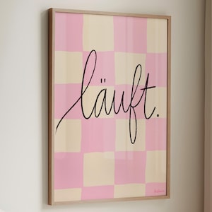 Könnte beinhalten: Ein gerahmter Kunstdruck mit einem rosa-cremefarbenen Schachbrettmuster. Der schwarze Schriftzug "läuft." ist mittig auf dem Druck platziert. Das Kunstwerk befindet sich in einem hellen Holzrahmen und hängt an einer weißen Wand.