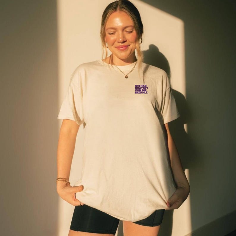 Puede incluir: Camiseta color crema con el texto "ICH HAB DIR DAVON ERZ&Auml;HLT" en morado. La camiseta tiene mangas cortas y un corte holgado. La persona de la imagen lleva pantalones cortos negros y un collar dorado.