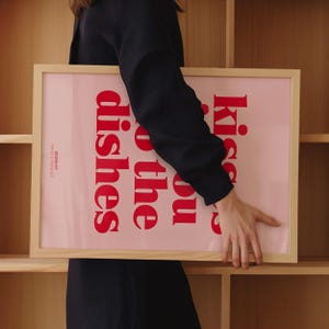 May include: A framed pink art print with the text "Kiss you o the dishes" in red. The frame is made of light-colored wood. The words "DESIGN BY FROST & FRECKLES" are printed in white on the bottom left corner.