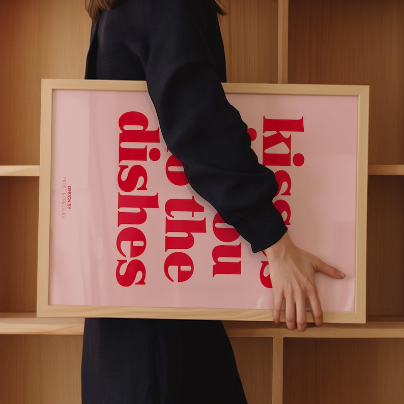 May include: A framed pink art print with the text "Kiss you o the dishes" in red. The frame is made of light-colored wood. The words "DESIGN BY FROST & FRECKLES" are printed in white on the bottom left corner.