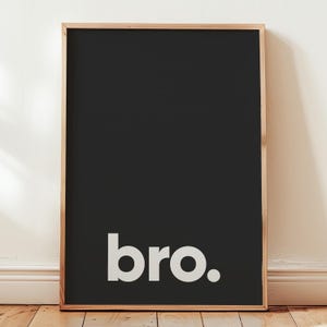 May include: A rectangular wall art print featuring a solid black background with the lowercase white text "bro." positioned towards the bottom. The print is encased in a light-toned wooden frame with a clean, simple edge and a flat profile.