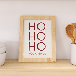 May include: A rectangular light-colored wooden frame containing a white print with the dark red text HO HO HO stacked vertically and the text HOL APEROL printed in smaller dark red letters at the bottom of the white background.