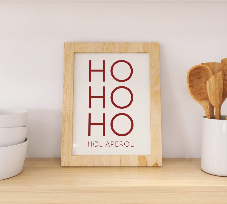 May include: A rectangular light-colored wooden frame containing a white print with the dark red text HO HO HO stacked vertically and the text HOL APEROL printed in smaller dark red letters at the bottom of the white background.