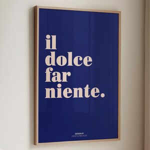 May include: A rectangular wall art print in a light wood frame featuring a deep blue background with the cream-colored text "il dolce far niente." centered in the upper portion and the smaller text "DESIGN BY FROST & FRECKLES" at the bottom.