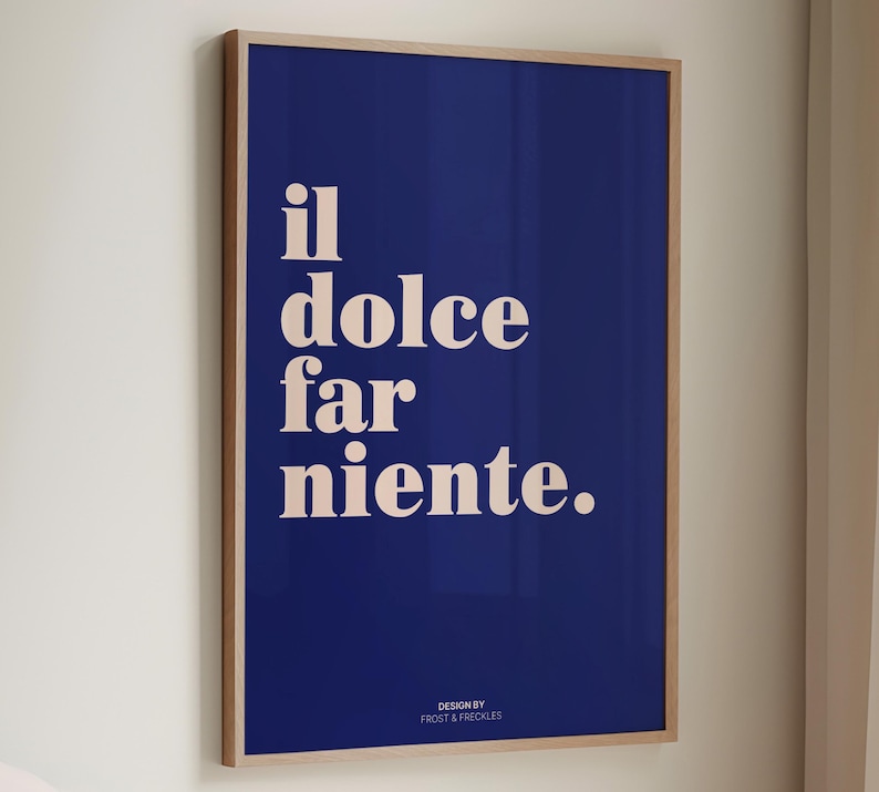 May include: A rectangular wall art print in a light wood frame featuring a deep blue background with the cream-colored text "il dolce far niente." centered in the upper portion and the smaller text "DESIGN BY FROST & FRECKLES" at the bottom.