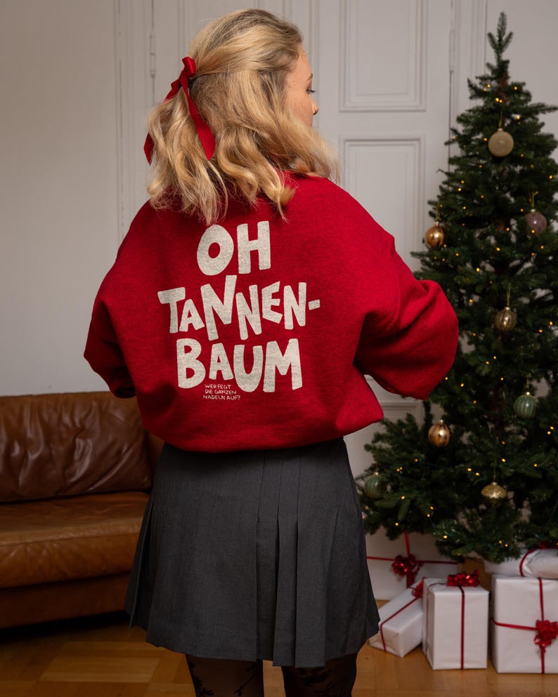 May include: A red long-sleeve sweatshirt featuring the white text OH TANNEN-BAUM and smaller text below that reads WER FEGT DIE GANZEN NADELN AUF? paired with a dark gray pleated skirt. The sweatshirt has a crew neckline and a relaxed fit.