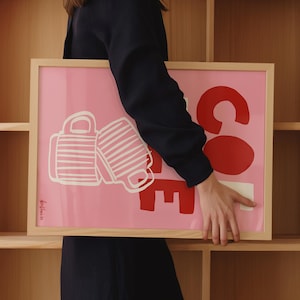 May include: Pink framed art print with the word "COFFEE" in red and white. The "CO" is large and round, with the "FFEE" stacked below. Two white coffee mugs are on the left side. The print is held by a person.