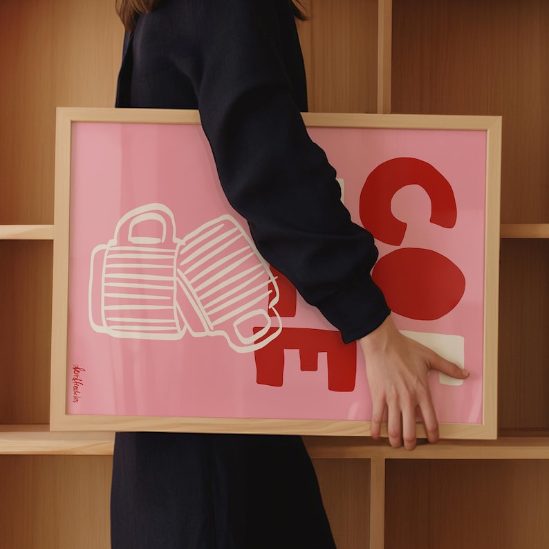 May include: Pink framed art print with the word "COFFEE" in red and white. The "CO" is large and round, with the "FFEE" stacked below. Two white coffee mugs are on the left side. The print is held by a person.
