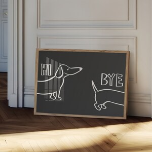 May include: A black framed print with a white outline of two dachshunds. The dachshund on the left says "HI" and the dachshund on the right says "BYE".