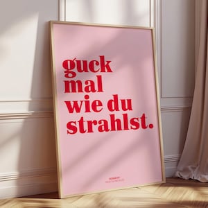 May include: A framed pink poster with red text that reads "guck mal wie du strahlst." The poster is in a light wooden frame and is leaning against a white wall. The words "DESIGN BY FROST & FRECKLES" are at the bottom.