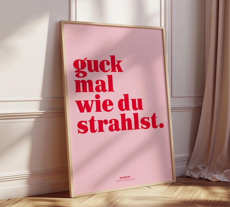 May include: A framed pink poster with red text that reads "guck mal wie du strahlst." The poster is in a light wooden frame and is leaning against a white wall. The words "DESIGN BY FROST & FRECKLES" are at the bottom.