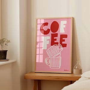 May include: A framed art print with the word "COFFEE" in red and white block letters on a pink background. Below the text are two stacked coffee mugs with white vertical stripes. The print is in a light wood frame.