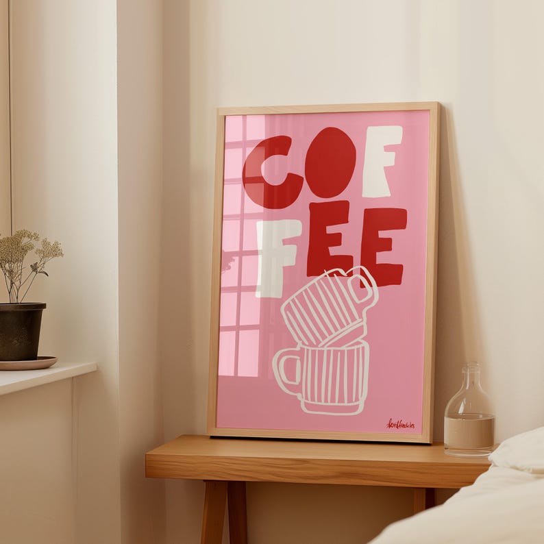 May include: A framed art print with the word "COFFEE" in red and white block letters on a pink background. Below the text are two stacked coffee mugs with white vertical stripes. The print is in a light wood frame.