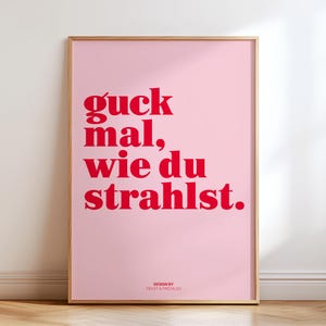 May include: A framed pink poster with red text that reads "guck mal, wie du strahlst." The poster has a wooden frame and the words "DESIGN BY FROST & FRECKLES" are at the bottom. The poster is leaning against a white wall.
