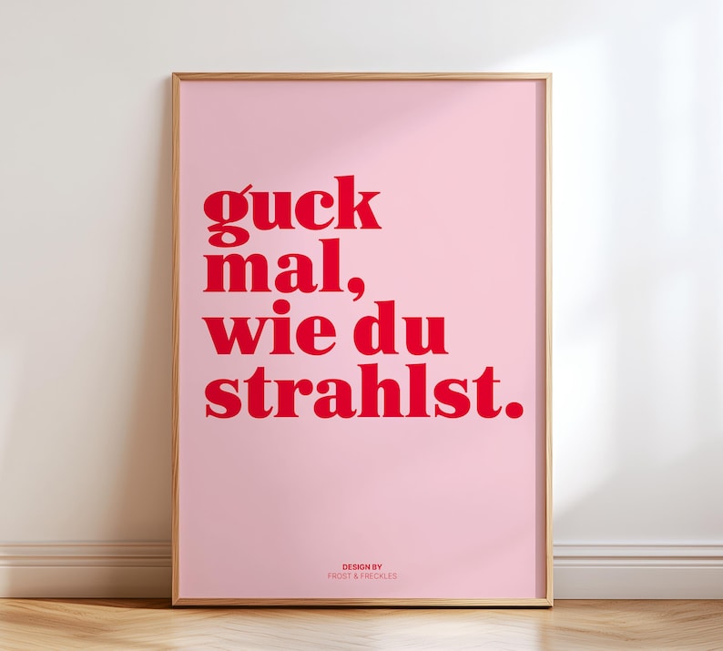 May include: A framed pink poster with red text that reads "guck mal, wie du strahlst." The poster has a wooden frame and the words "DESIGN BY FROST & FRECKLES" are at the bottom. The poster is leaning against a white wall.