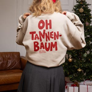 May include: A beige crewneck sweatshirt featuring the red text OH TANNEN-BAUM on the back, with smaller text below reading WER FEGT DIE GANZEN NADELN AUF? The sweatshirt is paired with a dark gray pleated skirt, and a red ribbon is tied in the hair.