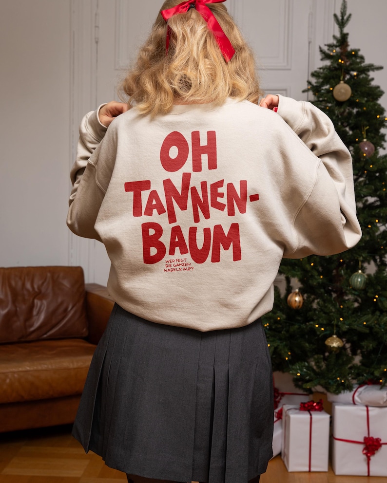 May include: A beige crewneck sweatshirt featuring the red text OH TANNEN-BAUM on the back, with smaller text below reading WER FEGT DIE GANZEN NADELN AUF? The sweatshirt is paired with a dark gray pleated skirt, and a red ribbon is tied in the hair.