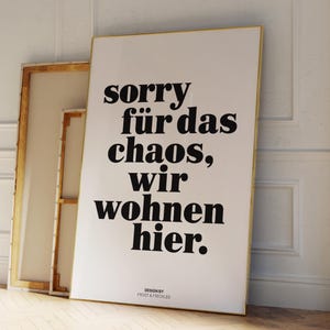 Könnte beinhalten: Ein weißes Poster mit schwarzem Text in einem goldenen Rahmen, mit der Aufschrift "sorry für das chaos, wir wohnen hier." Das Poster lehnt an einer weißen Wand. Dahinter ist eine weitere Leinwand zu sehen.