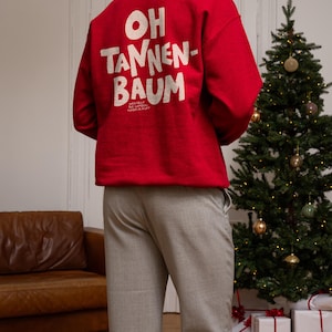 May include: A bright red long-sleeve sweatshirt featuring the white text OH TANNEN-BAUM printed on the back, with smaller text below reading WER FEGT DIE GANZEN NADELN AUF? The garment is paired with light gray trousers, creating a festive look.