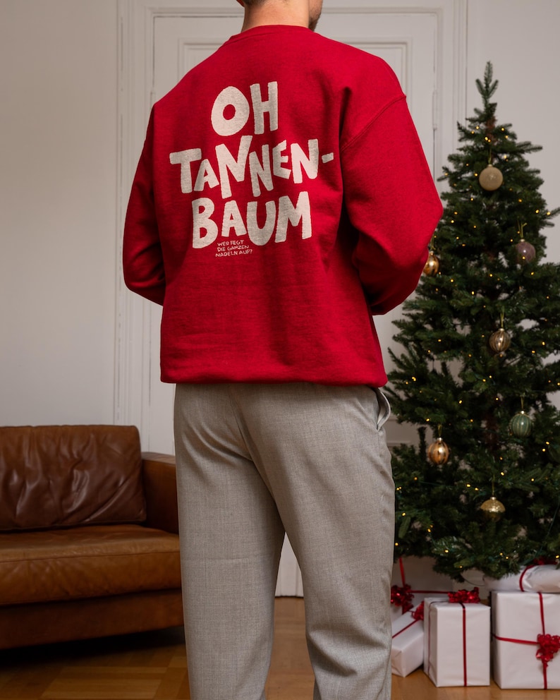May include: A bright red long-sleeve sweatshirt featuring the white text OH TANNEN-BAUM printed on the back, with smaller text below reading WER FEGT DIE GANZEN NADELN AUF? The garment is paired with light gray trousers, creating a festive look.