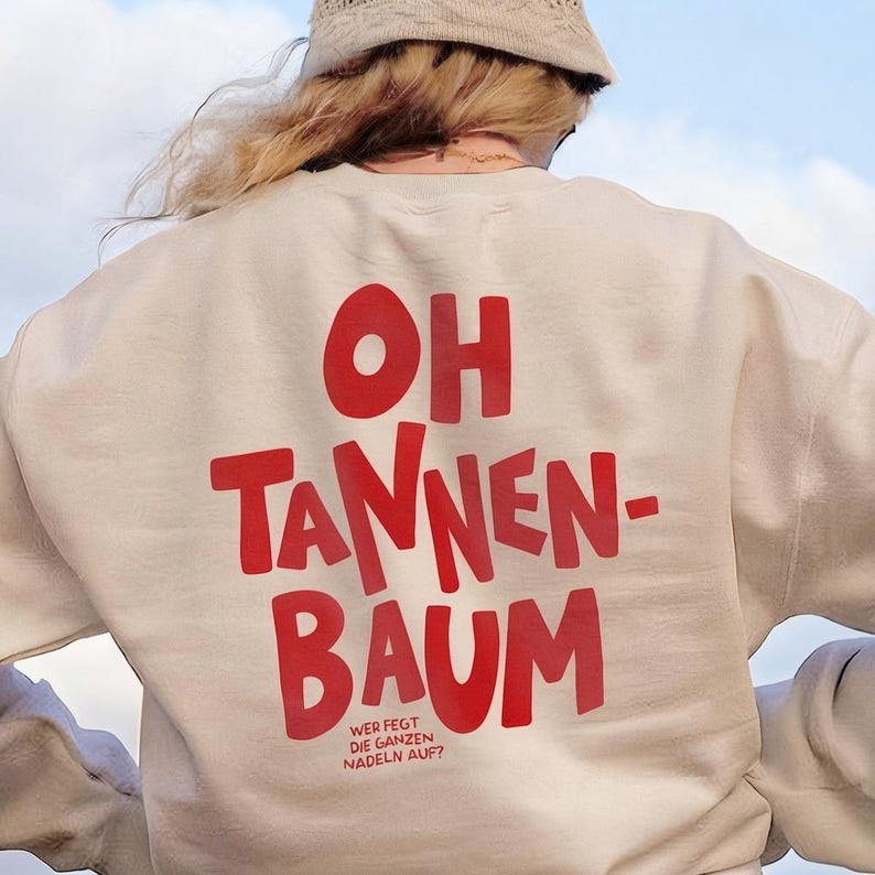 May include: A beige crewneck sweatshirt featuring large red text on the back that reads OH TANNEN-BAUM, with smaller red text underneath that reads WER FEGT DIE GANZEN NADELN AUF? The sweatshirt has long sleeves and a ribbed collar and cuffs.