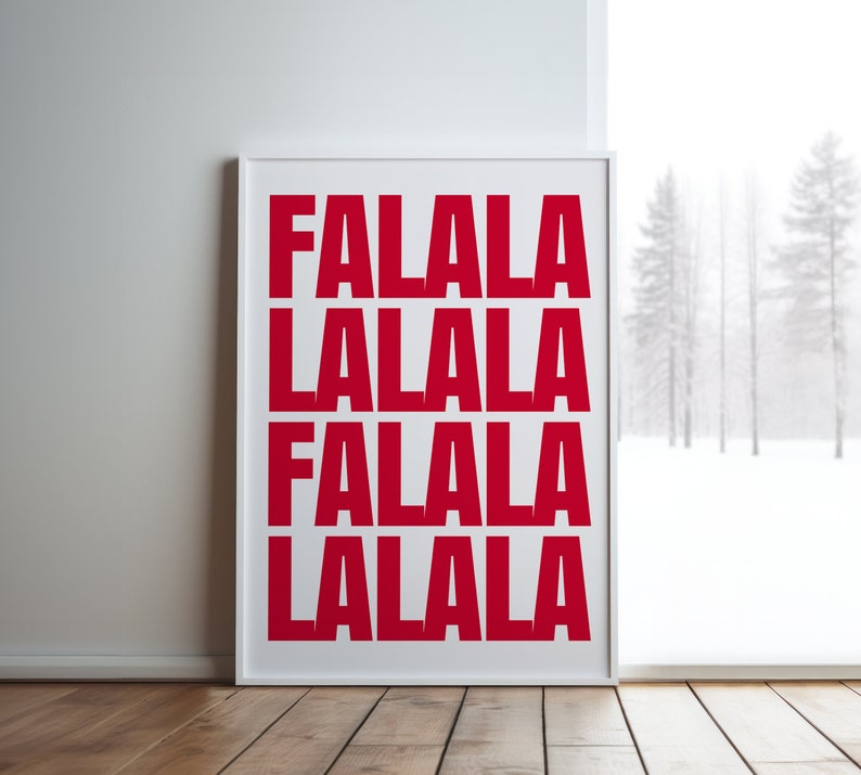 May include: A framed print with the words "FALALA" in bold red letters, repeated four times. The print is in a white frame and leans against a white wall on a wooden floor. A snowy winter scene is visible through a window.