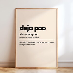 May include: A framed print with the words "deja poo" in bold black font. Below the title is the phonetic pronunciation and a German definition. The print has a light beige background and a wooden frame, suitable for wall decor.