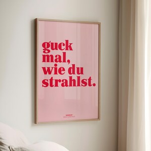May include: A framed pink poster with red text that reads "guck mal, wie du strahlst." The poster is in a light wood frame and is hanging on a white wall. The design is by Frost & Freckles.