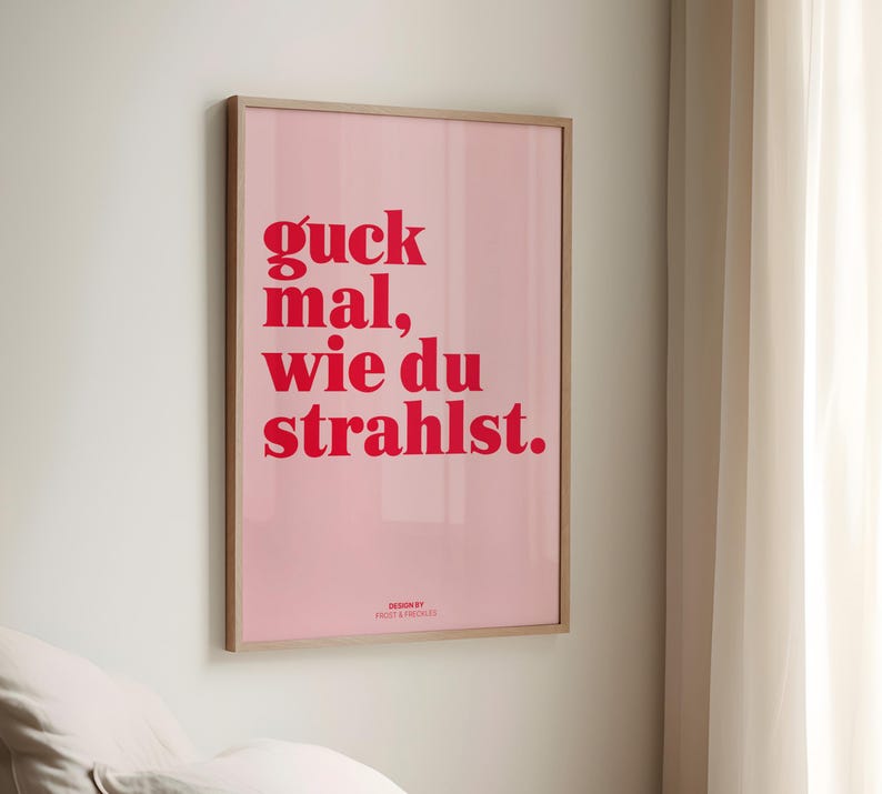 May include: A framed pink poster with red text that reads "guck mal, wie du strahlst." The poster is in a light wood frame and is hanging on a white wall. The design is by Frost & Freckles.