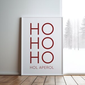 May include: A rectangular white framed wall art print featuring the dark red text HO HO HO stacked vertically, with the text HOL APEROL printed in smaller dark red capital letters underneath the main graphic on a white background.